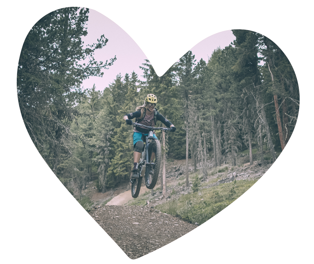 L'Almgasthof Geisler | è vicino alle piste ciclabili della Plose e permette di esplorare la Valle Isarco e la Plose, nonché Bressanone e l'Alto Adige in e-bike e mountain bike. L'immagine mostra una donna in sella alla sua bicicletta da downhill lungo uno stretto sentiero nel bosco. La donna salta una collina e in questo momento si trova in aria con la sua bicicletta. Intorno a lei ci sono alti abeti.