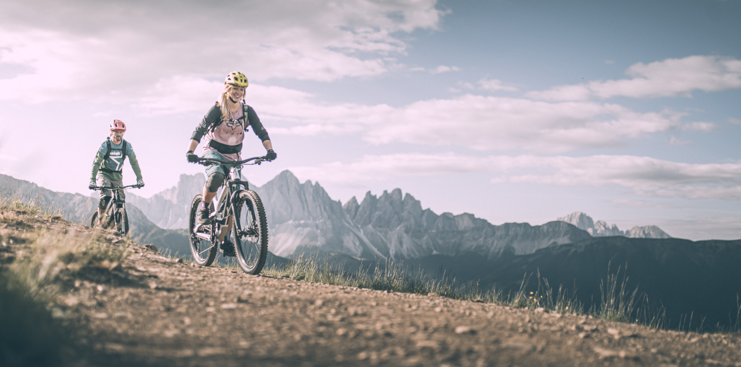 Almgasthof Geisler | vacanza direttamente in montagna e vicino alle migliori piste ciclabili dell'intera Valle Isarco L'immagine mostra due amici che percorrono uno dietro l'altro uno stretto sentiero in mountain bike sulla Plose. Stanno ridendo. Sopra di loro il cielo blu brillante e qualche nuvola, all'orizzonte troneggia il Geisler Villnöss.