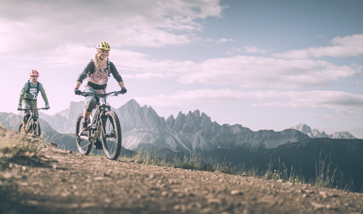 Almgasthof Geisler | vacanza direttamente in montagna e vicino alle migliori piste ciclabili dell'intera Valle Isarco L'immagine mostra due amici che percorrono uno dietro l'altro uno stretto sentiero in mountain bike sulla Plose. Stanno ridendo. Sopra di loro il cielo blu brillante e qualche nuvola, all'orizzonte troneggia il Geisler Villnöss.