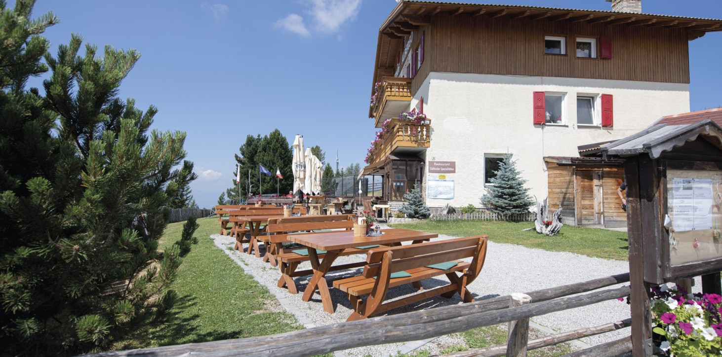 Almgasthof Geisler | das Beste aus Südtirols Speisekarte auf der barrierefreien Sonnenterrasse genießen Das Bild zeigt die barrierefreie Sonnenterrasse des Almgasthofes Geisler auf der Plose: Tische, eine Latschenkiefer, eine grüne Wiese