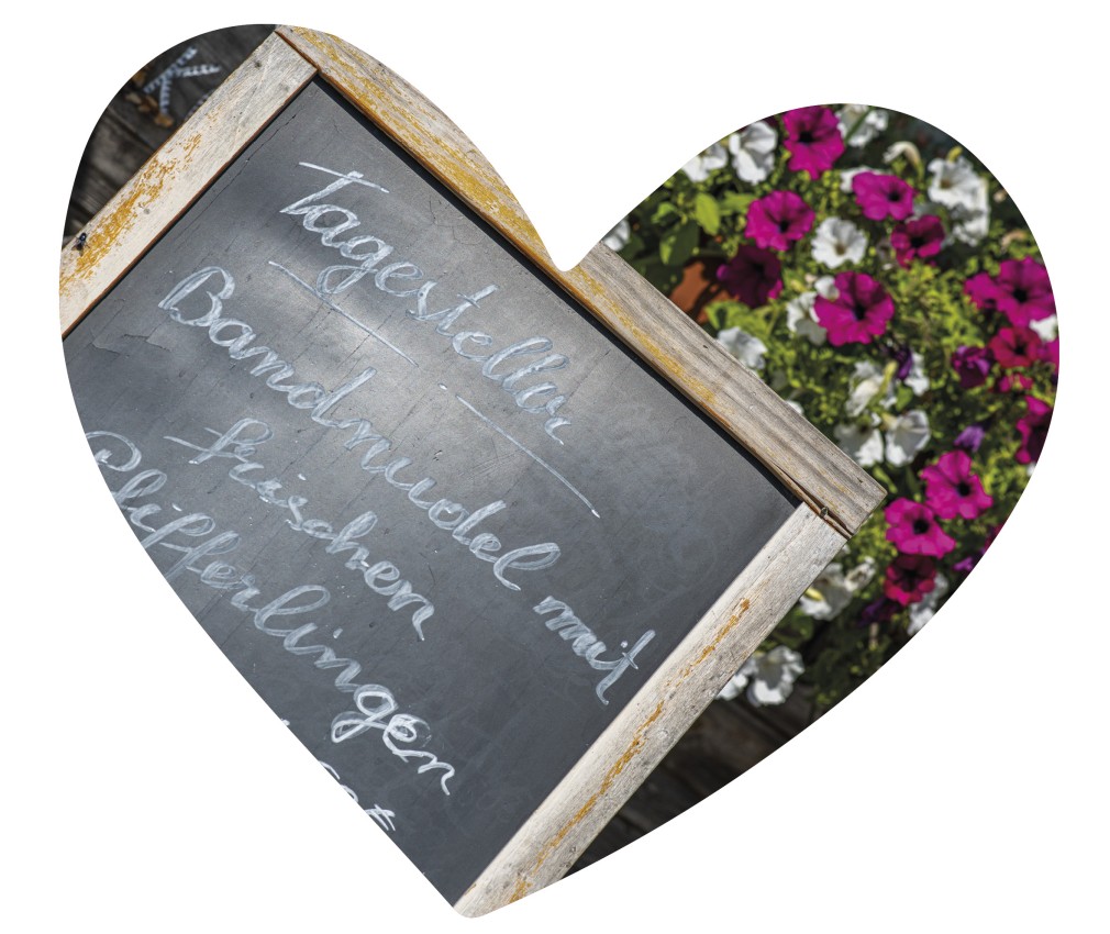 Almgasthof Geisler | traditionelle Südtiroler Gerichte und italienische Küche – die Abwechslung macht es spannend! Das Bild zeigt eine schwarze Tafel, auf der mit weißer Kreide die Tagesgerichte des Almgasthofes Geisler geschrieben worden sind. Im Hintergrund blühende Petunien.