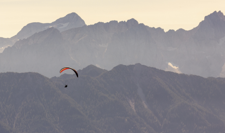 Almgasthof Geisler | Fare parapendio in ogni stagione significa godersi appieno la propria vacanza in Alto Adige in Valle Isarco L'immagine mostra un parapendio con un paracadute rosso che vola in aria sopra la Valle Isarco. Le Dolomiti più belle troneggiano all'orizzonte.