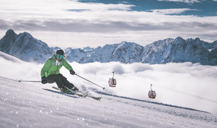 Almgasthof Geisler | Skiurlaub direkt neben der Piste auf der Plose im Südtiroler Eisacktal Das Bild zeigt einen Skifahrer auf der Plose. Im Hintergrund die Gipfel der Berge. Er trägt eine grüne warme Jacke und schaut in die Kamera.
