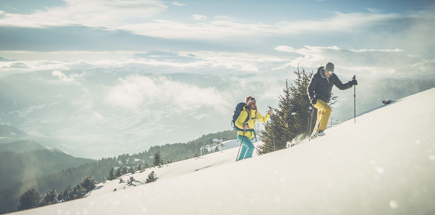 Das Bild zeigt zwei Menschen, wie sie durch eine tief verschneite Landschaft mit den Tourenskiern bergwärts wandern. Sie haben die Baumgrenze hinter sich gelassen und genießen einen fabelhaften Ausblick auf die umliegende Dolomiten–Bergwelt des Eisacktales.