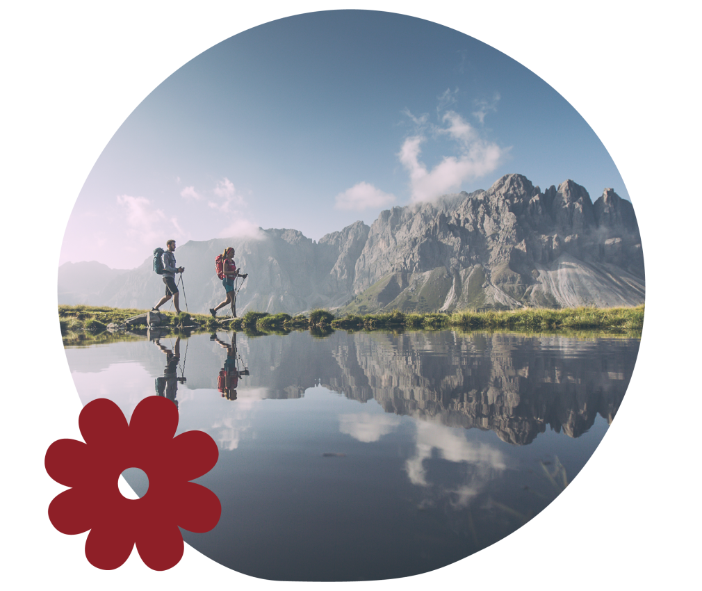 Almgasthof Geisler | escursioni sulla Plose e nelle Dolomiti: ecco cosa significa una vacanza sopra Bressanone, nella Valle Isarco in Alto Adige. Il quadro mostra una coppia che cammina lungo un lago di montagna. Sullo sfondo brillano le Dolomiti in splendide tonalità grigio chiaro. Al di sopra, brilla il cielo estivo di un azzurro intenso. Entrambi indossano zaini più grandi.