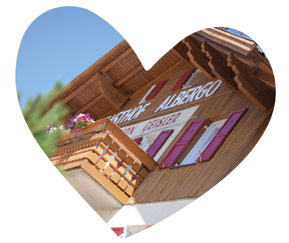 Almgasthof Geisler | Zimmer und Balkon für den Urlaub mitten in der Natur und direkt auf dem Berg über Brixen Das Bild zeigt das obere Stockwerk, einen Balkon und einige Fenster mit roten Jalousien. Darüber thront der Schriftzug Gasthof Albergo Pension Geisler. So wurde das Gasthaus zuerst genannt.